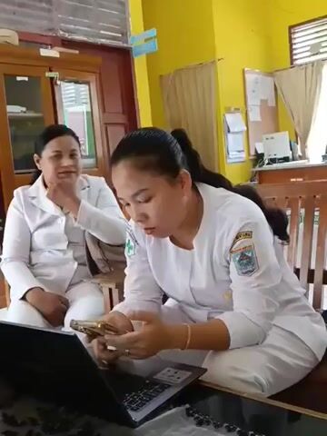 Two women are seated in what appears to be an office or classroom. The woman on the left is wearing a white uniform and has her hand near her mouth, as if thinking or speaking. The woman on the right is also wearing a white uniform and is looking intently at her mobile phone, which she holds in her hands. In front of her, a laptop is open on a desk. The room has yellow walls and wooden furniture, including cabinets and chairs. The overall impression is of a professional setting where the women are engaged in work-related activities.
