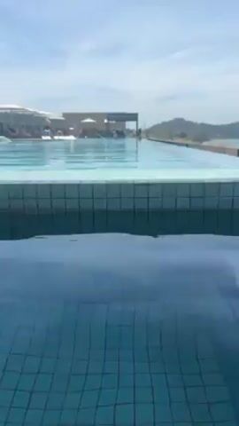 The video shows a view of an infinity pool during daylight. The camera moves slightly up and out, revealing a light blue tiled border in the foreground and the clear blue water in the background. The pool stretches towards the horizon where land and water meet under a bright, slightly cloudy sky. In the distance, people can be seen near lounge chairs and a covered structure, suggesting a resort or hotel setting.