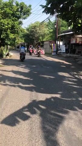 The video shows a dirt road with a few motorcycles. The first motorcycle is red and has two people on it. The second motorcycle is black and has a sidecar with a box on it. The third motorcycle is black and has one person on it. The motorcycles are moving away from the camera. The road is lined with trees and buildings. The sky is blue and there are some clouds.
