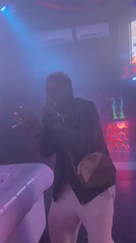 A Black man is singing into a microphone on a stage. He is wearing a black jacket, a white t-shirt, and white pants. He has a light-colored shoulder bag slung across his chest. He is gesturing with his hands. The stage is lit with colored lights that change from white to red and green, then to blue, and finally to orange. There is a white couch to the left of the stage and a wall with a honeycomb pattern behind him. There is smoke or fog in the air.