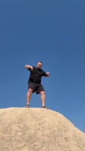 A man is captured in a low-angle shot, standing on top of a small, light-colored dirt or sand hill against a clear, vibrant blue sky. He is dressed in a black t-shirt, black shorts, and dark shoes. The man performs a series of movements, initially bending his arms at the elbows with his hands near his chest, then raising his fists upwards in a celebratory or energetic gesture, and finally lowering them again while looking out. He has a slight smile or smirk, suggesting he is enjoying the activity. The scene appears to be outdoors on a sunny day in Ürgüp, Turkey.