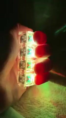 A hand is holding a stack of four transparent square objects that are lit up with different colors. The objects are stacked vertically. The colors of the light coming from the objects are blue, green, red, and green from top to bottom. The objects appear to be small and self-contained. The background is dark and out of focus, with a bit of what looks like a textured surface in the bottom left corner.