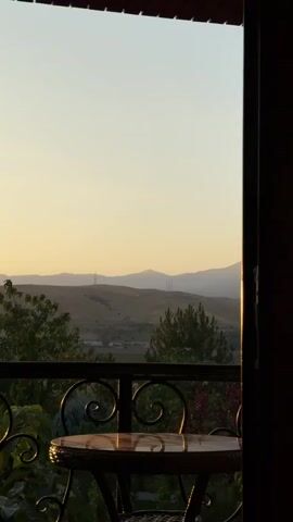 The video shows a balcony with a table and chairs. Beyond the balcony, there is a landscape with hills and mountains. The sky is a soft yellow, suggesting it is either sunrise or sunset. There are trees and some structures visible in the distance. The lighting is warm and diffused.