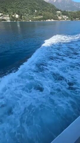 The video shows a view from a boat moving through the water. The boat is leaving a white foamy wake behind it. On the right side of the frame, there is a white bu oy attached to the boat by a rope. In the distance, there is a green, hilly coastline with trees and some buildings. The sky is clear and blue.