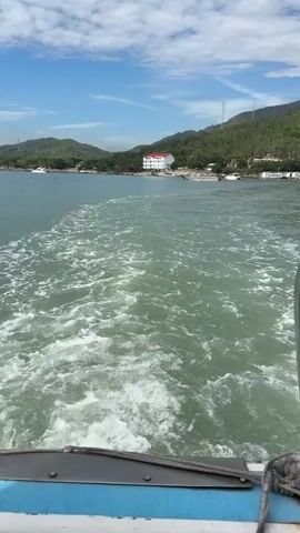 This is a POV shot from the back of a moving boat. The boat is leaving a white foamy wake behind it. The water is green and choppy. In the distance, there is a coastline with trees and hills. There is a white building with a red roof on the coastline, and some boats are moored in the water. The sky is blue with some white clouds.