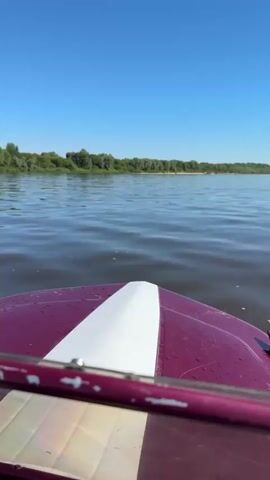 The video is filmed from the perspective of someone on a boat moving across a body of water. The bow of a dark pink and white boat is visible in the foreground. The water is rippled and appears dark blue under a clear, bright blue sky. In the distance, a treeline forms the horizon. The boat appears to be moving forward at a moderate speed. No other people or objects are clearly visible in the video. The overall impression is a sunny day on the water.