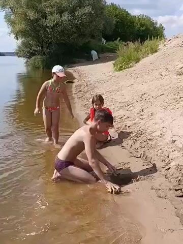 A group of three children are playing on a sandy riverbank. The water is shallow and calm.

In the foreground, a boy is kneeling in the shallow water, digging in the sand with his hands. He is wearing dark swim trunks. Behind him, a younger girl is also in the water, wearing a pink bikini and a white cap. She is looking at the boy. Next to her, another young girl in an orange life jacket is also playing with sand, leaning forward on her knees.

The riverbank is sandy and slopes upwards, with some dry grass and bushes on the higher ground. A large tree with green leaves is visible on the left side of the frame, its branches reaching over the water. In the distance, more trees and vegetation line the opposite bank of the river. The sky is partly cloudy, suggesting a warm, sunny day.

The children appear to be enjoying themselves, engaged in a typical summer activity of playing by the water. The scene evokes a sense of a relaxed, casual day spent outdoors. There is no visible text in the media. The location is identified as Nikiforovo, Russia.