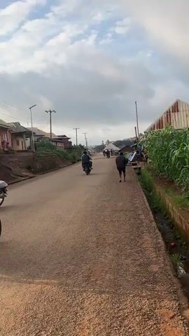 The video opens with a shot of a paved road in what appears to be a rural or semi-urban setting. The road is lined with buildings on the left and vegetation on the right. Several motorcycles are visible on the road, with people riding them. The sky is overcast with clouds.

In the foreground, a person is riding a motorcycle. They are wearing a green jacket and a black beanie. The motorcycle is white and appears to be covered with some plastic sheeting. The rider turns to look at the camera and smiles.

The camera then pans back to the road, showing other motorcycles in the distance. Some of the riders are wearing helmets. The buildings on the left appear to be simple structures, some with corrugated metal roofs. There are also power lines and utility poles along the road.

As the video progresses, the camera follows the road, showing more motorcycles and pedestrians. The overall impression is a busy street scene with a lot of motorcycle traffic. The environment looks somewhat undeveloped, with dirt or gravel visible on the sides of the road and in front of some of the buildings. The vegetation on the right side of the road is lush and green, suggesting a tropical or subtropical climate.