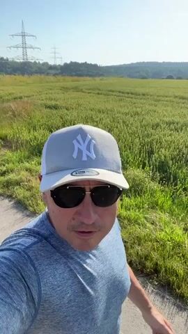 A man wearing a grey and white baseball cap with a white NY logo and sunglasses is walking in what appears to be a field of wheat or tall grass. He is also wearing a blue t-shirt and is looking at the camera. The sun is shining, and the sky is clear. In the background, there are trees and a power line tower. The man is on a dirt path.