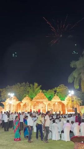 At night, a large gathering of people, mostly men in white shirts, are at an outdoor event with a brightly lit, ornate structure in the background. The structure appears to be a stage or backdrop, decorated with intricate designs and illuminated with orange and green lights. Fireworks are visible in the night sky. People are milling around and walking casually. The overall atmosphere is celebratory and festive.