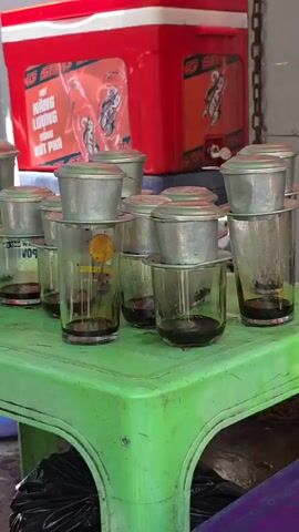 A medium shot shows a collection of Vietnamese coffee filters, each with a filter in place and a glass beneath it, resting on a bright green plastic table. Behind the table, a large red cooler with graphics is visible. The coffee filters appear to be metal and are stacked in multiple rows, suggesting preparation for making several cups of coffee. The glasses underneath are partially filled with a dark liquid, presumably coffee. The environment appears to be outdoors or in an open-air setting, given the lighting and the presence of the cooler.
