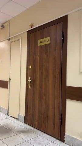The video shows a close-up view of a wooden door with a gold plaque that reads "ПРИЁМНАЯ" (Reception). Above the door, on the ceiling molding, there is a small icon of a religious figure. The camera then pans to the left, showing a hallway with beige walls, a white tiled floor, and a dark wood trim running along the walls. Several doors are visible along the hallway, as well as some benches. Further down the hallway, a person in a white shirt and dark pants is walking away from the camera. The hallway is illuminated by fluorescent ceiling lights.