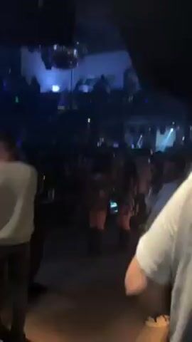 The video shows a crowded nightclub with green and blue lighting illuminating the dancing people. A disco ball hangs from the ceiling. People are standing, walking, and dancing. The camera moves slightly, showing different areas of the club and people.
