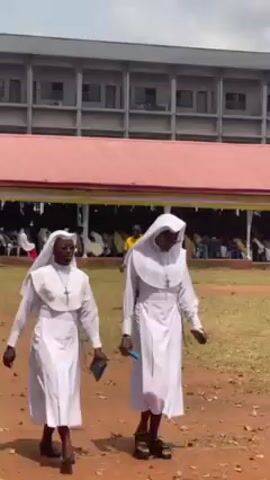 Two individuals dressed as Catholic nuns are walking across a grassy area with a reddish dirt path. They are wearing white habits, white veils, silver cross necklaces, and dark sandals. One is holding a small dark object in their left hand and the other is holding a blue object. Behind them is a building with a red roof and multiple windows. In the background, there is a white tent-like structure with people seated under it. There are also a few individuals wearing uniforms or security attire in the background.