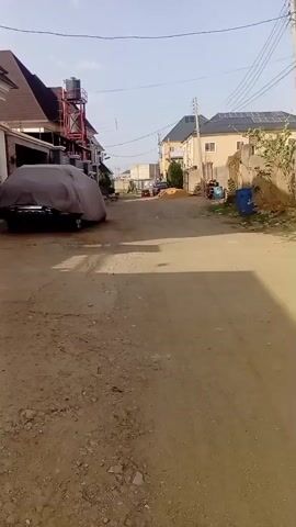 The video shows a dirt road in a residential area. On the left side of the road is a house with a car covered by a gray tarp. A water tower is attached to the house. On the right side of the road, there are some blue trash cans, a pile of dirt, and a few houses. Power lines are strung across the street. The sky is clear. There is no visible human activity.
