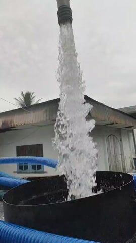 A video captures water gushing from a hose into a black container. The hose is positioned above the container, with water spraying downwards in a powerful stream. The scene is set outdoors, with a building in the background. The building appears to be constructed with corrugated metal roofing and white walls. Blue hoses are visible on the ground, coiled around the black container. The sky is overcast, with a visible sun in the upper right portion of the frame. A palm tree is visible to the left of the building. The overall impression is one of industrial or agricultural activity.