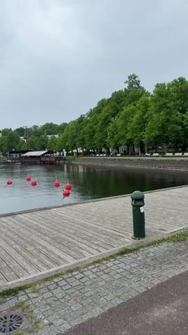 The video features a handheld shot on a cloudy day, starting with a view of boats docked on the water and moving right to showcase a large ship converted into a restaurant or bar. The ship has an outdoor seating area with patrons, umbrellas, and flower boxes, and a tall smokestack. A wooden dock is visible in the foreground, with paved ground and a drain cover. Trees line the far bank, and buildings are visible in the background.