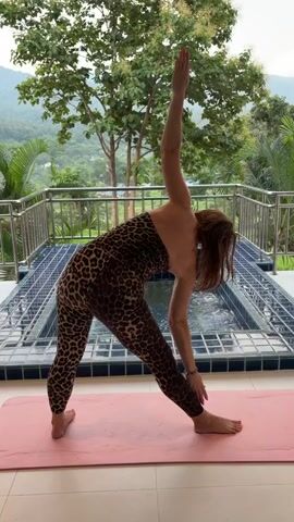 A woman with reddish-brown hair, wearing a leopard-print bodysuit, performs a yoga pose on a pink mat outdoors. She stands with her legs wide apart, one foot forward and slightly angled, the other foot turned out perpendicular to the first. Her torso is bent forward, with her hands clasped behind her back. Her head is tilted back, and she appears to be looking upward. In the background, there is a swimming pool with bubbling water, a metal railing, lush green trees, and a cloudy sky. The lighting suggests it is daytime.