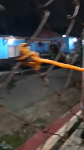A close-up shot of an orange rope tied to a rusty metal fence. In the background, there is a blurry building with blue walls and illuminated windows. The scene appears to be outdoors at night.
