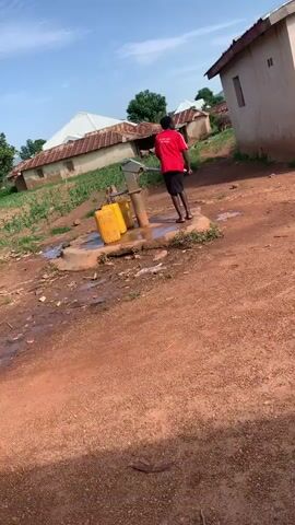 A person wearing a red t-shirt and dark shorts is operating a manual water pump. Two yellow water containers are positioned next to the pump, one filled with water. The pump is located on a concrete platform, which is surrounded by dirt and puddles of water. In the background, there are several small houses with corrugated metal roofs, and some vegetation. The sky is blue with some clouds.