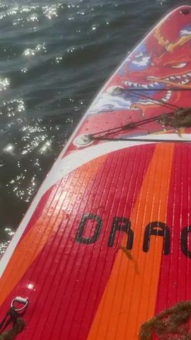 A first-person perspective video shows the front of a paddleboard with a red and orange striped design and a red dragon graphic on its surface. The paddleboard is on a body of water with moderate waves and sunlight reflecting off the surface. The sky is blue with scattered white clouds. The paddleboard is moving forward through the water.