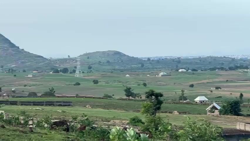 A wide drone shot pans across a landscape. In the foreground, several small, partially constructed buildings are visible, along with a low concrete wall. In the middle ground, there are scattered houses with colorful roofs, some with dome-like structures, and green fields. Hills and more distant buildings can be seen in the background. A small airplane is visible in the sky. The overall scene depicts a rural or semi-rural area with scattered development.