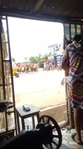 The video shows a group of children in yellow and blue school uniforms standing on a street, near a yellow auto rickshaw and a few motorcycles. The children are accompanied by at least one adult. Across the street, a sign for "Sky Gold Hotel and Suite" is visible. The video appears to be taken from inside a shop, where a sewing machine and a small table are present.