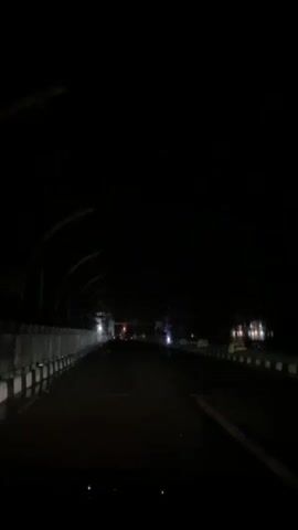 A dark, narrow road curves under a covered walkway. Streetlights cast dim, yellow glows in the distance. A motorcycle with its headlight on speeds past, followed by a truck.
