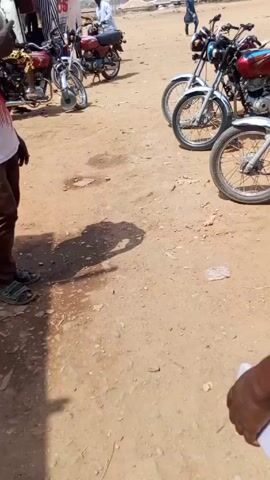 The video depicts a sunny day in what appears to be a market or transportation hub. Several motorcycles are parked on a dirt ground, showing signs of heavy foot traffic and general outdoor use. 

In the opening scene, the focus is on the shadow of the person filming. Subsequently, the videographer, who is wearing a light-colored Liverpool jersey, walks towards someone with braided hair. The camera perspective shifts, briefly showing a man sitting on a red motorcycle next to a makeshift wooden stall selling clothes under an umbrella. The man on the motorcycle looks directly at the camera and seems to be waiting for someone. The camera then focuses back on the ground near the motorcycle, showing the dusty dirt surface.