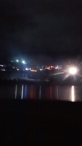 The video is a shaky, nighttime shot of a brightly lit vehicle moving towards the viewer. The vehicle's headlights are intensely bright, creating a starburst effect and washing out much of the surrounding scene. In the background, there are distant lights, suggesting a town or city. A bridge structure is faintly visible in the middle ground. The foreground is dark, with reflections of colored lights, likely from the vehicle or surrounding environment, visible on what appears to be a body of water. The overall impression is one of movement and strong artificial light against a dark backdrop.