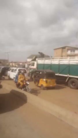 The video shows a busy street in what appears to be an African city. There's a large truck stopped with a man on top, seemingly adjusting or securing cargo. A yellow and black auto-rickshaw is nearby. People are riding motorcycles, and a white vehicle is also visible. The sky is overcast with some patches of blue. The road is dusty, and there are buildings visible in the background. The camera appears to be inside a moving auto-rickshaw, offering a first-person perspective of the street scene.