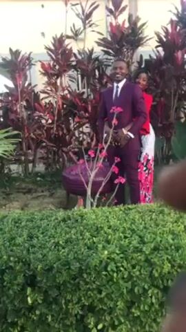 A person records a man and woman posing for a photo outside. The man is wearing a plum suit and tie with a white dress shirt. The woman is wearing a red top and a white skirt with red floral designs. The woman has her arms wrapped around the man’s midsection. The couple is standing in a garden with red plants behind them. In the foreground is a trimmed green bush, and in front of that is a small shrub with pink flowers. There is a purple, round garden stool next to the pink flower shrub. In the upper portion of the shot, the corner of a light-colored building is visible.