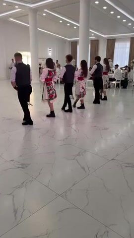 A group of six young people, four women and two men, are dancing in a large, well-lit hall with a high ceiling and marble-patterned floor. They are all dressed in traditional folk costumes. The men wear black vests over white shirts and black pants. The women wear white dresses with colorful embroidered patterns and black boots. The group is arranged in a line, facing the same direction, and appears to be performing a choreographed dance. In the background, several other people are seated at tables, and some individuals are standing and observing the dancers. The overall atmosphere suggests a formal event or celebration.