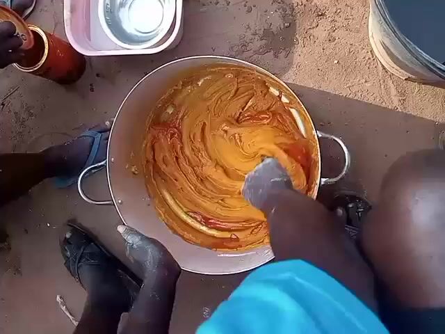 The video shows a person using their hand to mix a large pot of what appears to be a yellow and red paste. The pot is silver and has two handles. The person's hand is covered in a white powder.  The person is wearing a light blue shirt.  Another person's legs are visible in the background, wearing blue flip flops and black sandals. A jar and a pink bucket with small bowls inside sit to the left of the pot. The ground is a reddish-brown dirt or sand. The video is shot from a top-down perspective.