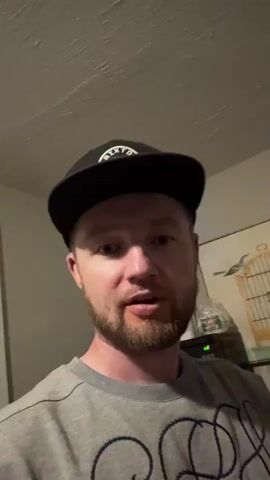 A man wearing a black baseball cap and a grey t-shirt looks into the camera and holds up two fingers in a peace sign. He has a beard and appears to be speaking. The background is out of focus but shows a ceiling, a wall, and some objects on a shelf including a bottle and what looks like a birdcage.