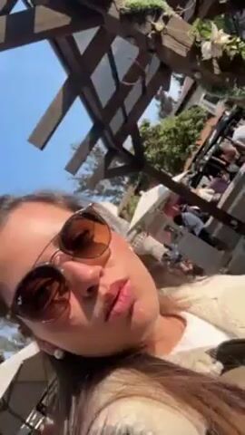 A woman with sunglasses is outdoors, taking a video of herself. The camera pans up to show a stone tower, and then back down to the woman's face. There is a wooden structure above her, and people and buildings in the background. The day is sunny.