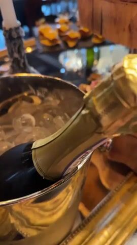 The video shows a close-up of a silver ice bucket with a champagne bottle nestled inside. The bucket is filled with ice cubes. Beside the bucket is a two-tiered display of appetizers: small, open-faced sandwiches topped with various ingredients like salmon, meat, and vegetables. A tall, decorative candle holder with a lit white candle stands next to the food display. In the background, a dining table is set for a formal meal with wine glasses and plates. The scene suggests a celebratory event or gathering. The lighting is warm and inviting.