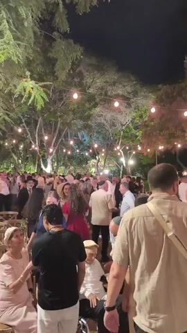 A large gathering of people is seen at night, outdoors, possibly at a wedding or celebration. String lights are hung in trees, illuminating the scene. Many people are dressed in formal or semi-formal attire. Some individuals are dancing, while others are standing and conversing. A man in a wheelchair is visible. The overall atmosphere appears festive.