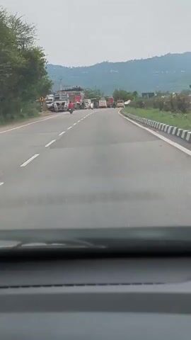 The video shows a view from inside a vehicle looking out at a two-lane road. There is a line of trucks and other vehicles stopped on the side of the road, indicating a traffic jam or obstruction. A person on a scooter is passing by the stopped vehicles. In the background, there are trees and hills under a cloudy sky. The road has white lines marking the lanes and a barrier on the right side.