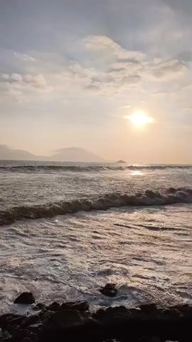 The video shows a view of the ocean at sunset. The sun is low in the sky and appears as a bright orb surrounded by a halo of light. Clouds are scattered across the sky, and their undersides are illuminated by the setting sun. Waves are crashing on the shore, and the water is a mixture of blues, grays, and whites. In the distance, there are mountains, their silhouettes softened by the haze of the setting sun. The foreground of the image is a rocky shoreline, dark and wet from the ocean spray. The overall impression is one of natural beauty and tranquility.
