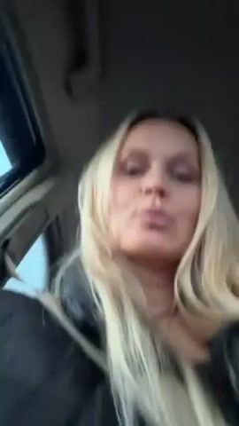 The video shows a blonde woman sitting in the driver's seat of a car. She is wearing a black jacket and a seatbelt. She is looking at the camera and making a kissy face. She runs her fingers through her hair. There is a backseat visible. Music is playing in the background.