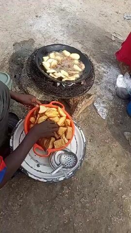 The scene is an outdoor cooking setup on what appears to be a concrete or similar rough surface. A person is sitting on the ground near the cooking area. To their left, there’s a green bucket. At the center, there’s a large white tray on which sits an orange colander filled with fried food items, some cut in the shape of apple slices and others appearing as square pieces. Next to the colander, on the tray, is a slotted spoon. Above this tray, there’s a frying pan with more of the apple-slice shaped food items frying in oil. The pan is placed on what looks like a wire grate, supported by stones, possibly serving as a makeshift stove. To the right of the pan, there are some plastic bags and another person in a red garment. The person on the ground is using their hands to sort through the food in the orange colander.