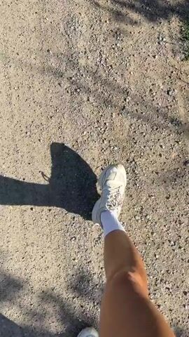 The video shows a person walking on a dirt path. The camera is positioned at eye level and looking down at the person's legs and feet. The person is wearing white sneakers and white socks. The path is covered in gravel and dirt, and there are shadows cast by trees on the ground. The lighting suggests it is daytime. The person's legs are tanned. The video appears to be shot from the perspective of the person walking.