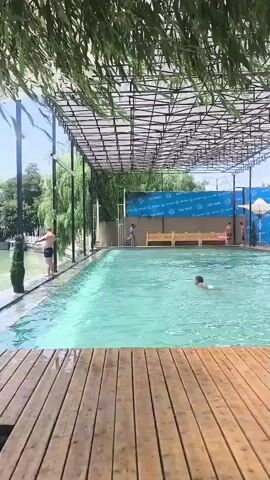 A video clip shows a rectangular swimming pool with turquoise water. The pool is covered by a metal frame structure with a dark mesh roof, providing shade. Wooden decking surrounds the pool. Several people are visible in and around the pool. On the left side of the pool, a person stands on the edge, facing away from the camera. In the water, one person swims towards the right. Further back in the pool, several other individuals are present. Along the far edge of the pool, there is a row of seating areas and some buildings in the background. Green foliage hangs down from above, framing the top of the scene. The overall environment appears to be an outdoor recreational area on a sunny day.