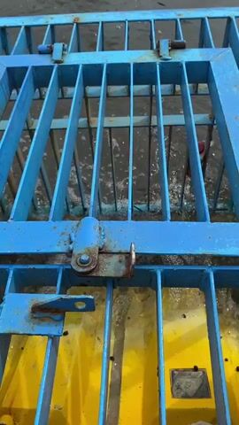 A blue metal grate with rusty hinges is positioned over a yellow surface that is partially submerged in water. The water is choppy and appears to be flowing. Through the grate, a red object is visible in the water.