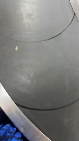 The video shows a close-up view of a black conveyor belt, likely in an airport baggage claim area. The belt is wide and appears to be moving. There are some smudges and scratches on the surface of the belt, as well as a few small, light-colored specks. In the lower left corner, a portion of dark fabric with a blue pattern is visible, suggesting it may be part of someone's clothing or luggage. A shiny metal railing runs along the bottom edge of the frame. The overall environment appears to be indoors, under artificial lighting, given the lack of shadows and the slightly grainy quality of the footage. The video focuses on the surface of the conveyor belt as it moves, with occasional glimpses of the surrounding metallic structure.