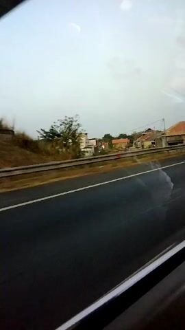 The video shows a view from inside a vehicle, looking out a window at a highway and a rural landscape. The highway is paved and has lane markings. A metal guardrail separates the highway from the side of the road. On the other side of the guardrail, there is dry grass and some trees. In the distance, there are a few houses with red tile roofs. The sky is overcast and light. The camera is moving, indicating that the vehicle is in motion. There are no people or animals visible in the video. The overall impression is of a typical roadside view while traveling.