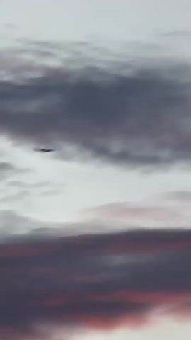 This vertical video captures a silhouette of an airplane flying against a backdrop of dramatic clouds at dusk. The sky is a canvas of grey, purple, and vibrant pink hues, indicating sunset or sunrise. The clouds are textured and layered, with some appearing wispy and others more dense. The airplane, a dark silhouette, is positioned in the upper left quadrant of the frame, appearing to fly horizontally. The overall mood is serene and somewhat majestic, evoking a sense of travel and the vastness of the sky. There is no visible text in the media. The location context provided is Khan Mean Chey, Cambodia.