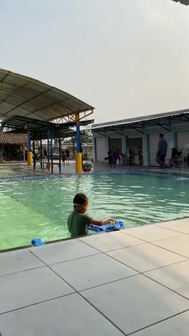 A vertical video captures a boy in a green shirt and goggles standing in a shallow swimming pool. He is holding a blue floatation device with a yellow handle. The pool water is green and appears to be clear. To the left of the boy, under a covered area with a yellow roof and blue support beams, other people are visible, including a child in a yellow shirt and a person in a dark shirt. To the right of the pool is a building with a white facade and a dark roof, housing several stalls or service areas. People are gathered in front of this building, including a person in a blue shirt and dark pants, and a few other individuals. A fountain with water spouting is visible on the right side of the pool. The ground around the pool is tiled with white and light grey squares. The sky is overcast. The video appears to be filmed during daylight.