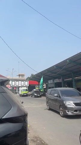The video shows a street scene on a sunny day. In the foreground, a dark-colored car is parked on the left side of the road. To the right, a scooter carries two people, a male driver and a female passenger, both facing away from the camera and moving forward. They are dressed casually; the passenger wears a hijab. Next to the scooter, a silver SUV is also moving forward. Beyond the vehicles, a long, covered market structure with a metal roof is visible, adorned with Indonesian flags. The structure appears to be lined with stalls. A large green flag is also visible hanging from the structure. Further in the background, several other vehicles, including a lime green truck, are on the road, and some buildings are present. The sky is clear and blue. The overall environment suggests a bustling street scene in a market area.